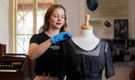 A person stands in Claremont Museum tending to a dress on a mannequin. There is a piano-style instrument, signage and a window in the background.