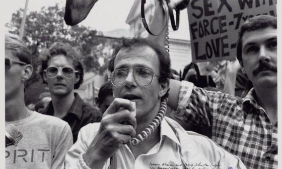 A black and white image of a group of mean demonstrating. The man front and centre is holding a microphone to his mouth, while the man to the right is holding a megaphone speaker up in the air. There are placards and flags in the air in the background.
