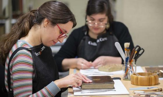 Two people work at a table with tools and brushes, restoring old books. Both wear aprons. The person in the foreground has long brown hair and red glasses, focusing on a book. The other person is slightly out of focus in the background.