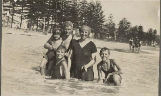 A black-and-white photo of a smiling family in swimsuits, standing together in shallow water at Manly Beach. The background shows pine trees and other beachgoers. The picture was taken in 1923 in New South Wales, Australia.