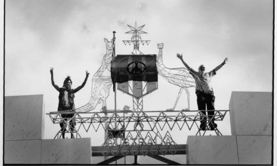 Two demonstrators stand on a structure at Parliament House, Canberra, raising their arms in the air beside an Australian Coat of Arms sculpture. A large peace flag is draped over the emblem. The black-and-white image captures the scene from below, with a cloudy sky in the background.