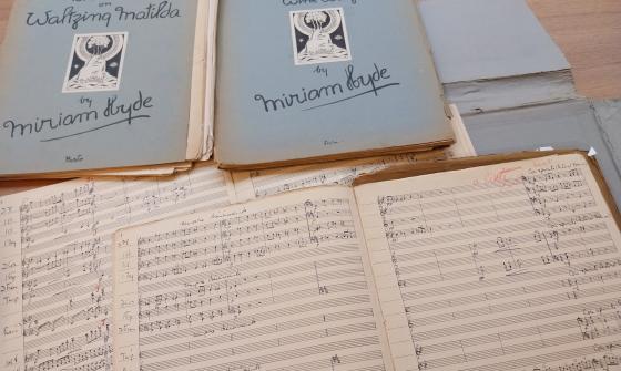 Sheet music books, some open and some closed, spread over a wooden table. The closed books' covers read 'Fantasia on Waltzing Matilda by Miriam Hyde' and 'Orange Growing and Wine Waltz by Miriam Hyde'