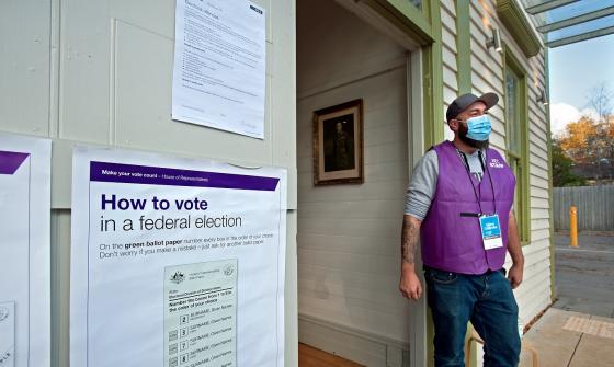 Man wearing face mask and purple vest standing new a doorway. On the wall around the door are instructions on how to vote in a federal election.