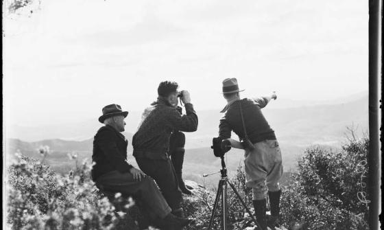 Three men at the peak of a mountain looking at the view with binoculars and pointing