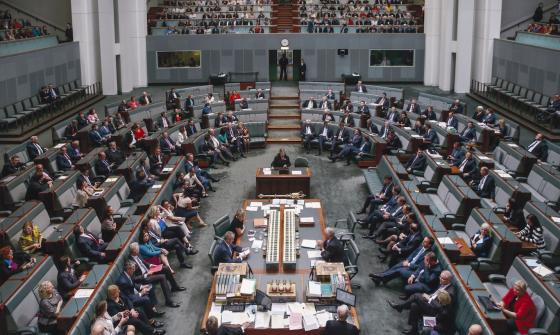 View of the Australian House of Representatives in Parliament House from slightly above. Members of the House of Representatives sit throughout the room, and members of the public sit in allocated areas