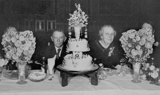 Husband and wife sitting at a table with a large cake and vases of flowers in front of them