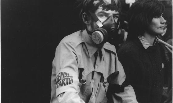 Young man, maybe late teens, wearing a grey shirt with a 'Friends of the Earth' armband wearing a gas mask
