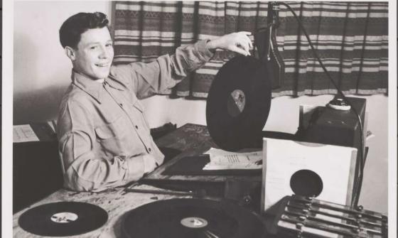 At the left of the photo, a young man with short dark hair is sitting down at a radio desk, smiling out at the camera and with his left hand he balances a record on its edge on the desk.