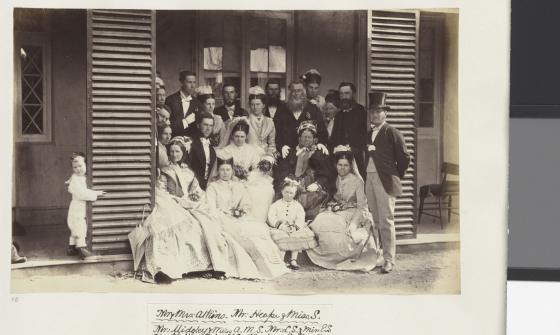 A black-and-white photograph showing a large wedding party posed on the steps of a wooden house. The group includes men, women, and children dressed in formal Victorian attire. The women are in elaborate dresses, some holding small bouquets. The men wear suits, some with top hats, and a small boy peeks around a door frame to the left.