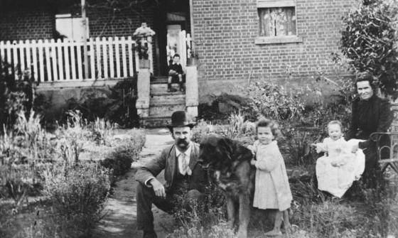 A black-and-white photo of a family posed outside a brick house with a white picket fence. In the foreground, a man in a bowler hat sits beside a large dog, while a young girl stands nearby. A seated woman holds a baby on her lap, and a child is visible on the steps of the house in the background. The garden surrounding them is filled with overgrown plants.
