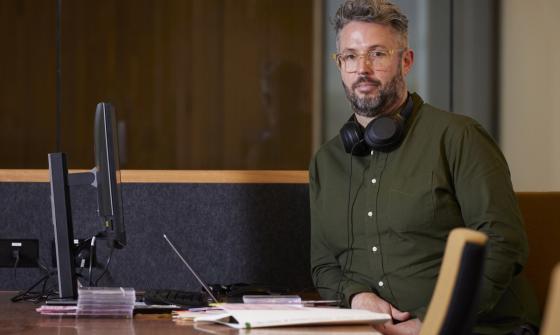 A man in glasses and a green shirt with headphones around his neck is sitting at a desk in front of a computer.