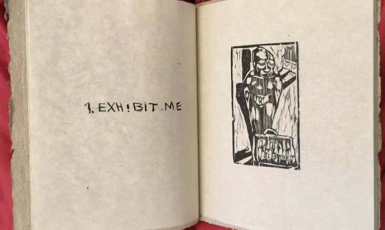 A large book sitting propped open on a red piece of material. The left hand page shows text that reads '1. Exhibit me' and the right hand page shows a stamped black ink image.