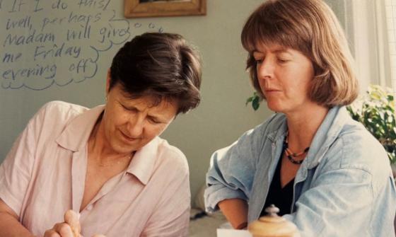 Two women looking down at something out of frame. One is wearing a pink shirt and one is wearing a blue shirt.