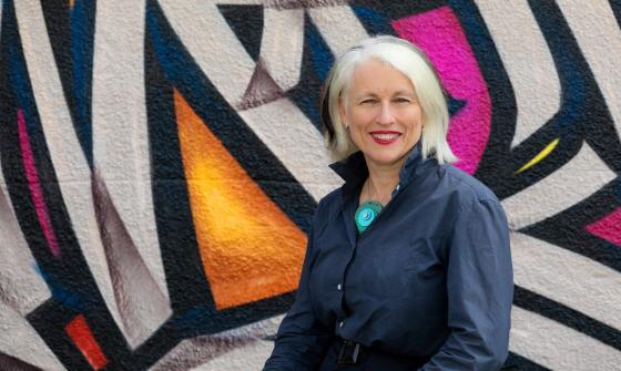 A portrait image of Genevieve Jacobs AM standing in front of a colourful wall