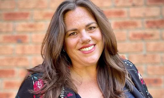 A headshot of a smiling woman with long, brown hair. She is wearing a colourful floral jack and is standing in front of a brick wall.