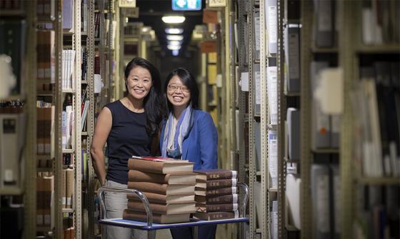 Two women standing behind a trolley with books on it in the Library stacks.