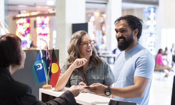 Man and woman smiling as they are being greeted and having questions answered at the front information desk in the Library foyer