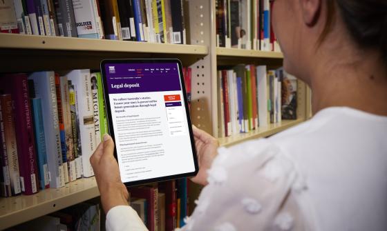 Woman standing in front of a large bookshelf holding up a tablet showing the 'Legal Deposit' page of the National Library website