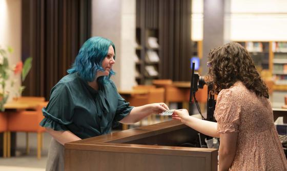 A person with teal coloured hair stands at the Library’s wooden service desk, handing an item to another person across the desk. The background shows the Library interior with bookshelves and tables.