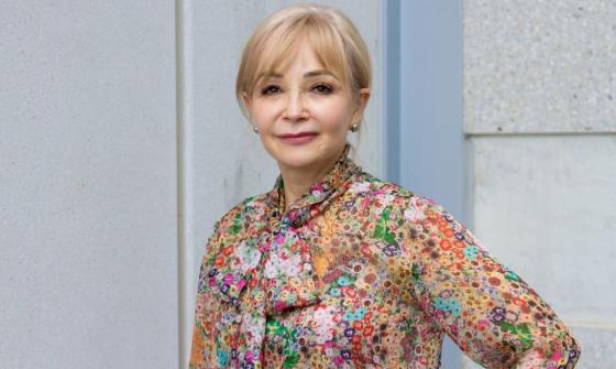 Portrait photograph of a woman with blonde hair, wearing a colourful floral shirt and with one hand on her hip.