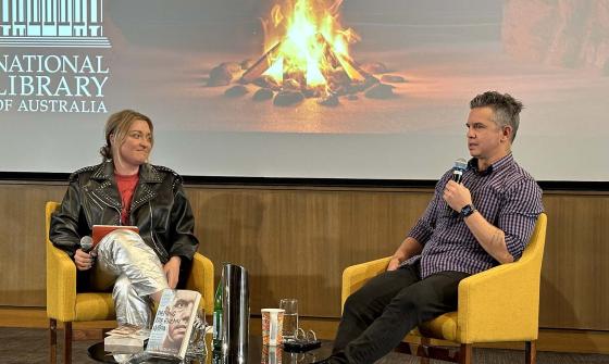 A man and a woman sit in chairs on stage with a picture of a campfire behind them. The woman holds a microphone and the man is speaking into a microphone.