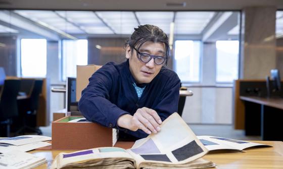 2023 National Library of Australia Fellow Dr Tets Kimura sits at a library desk examining a large open book filled with swatches of fabric or coloured paper. They have dark hair and wear glasses and a navy blue shirt. Next to them on the desk is a wooden box with more books in it.
