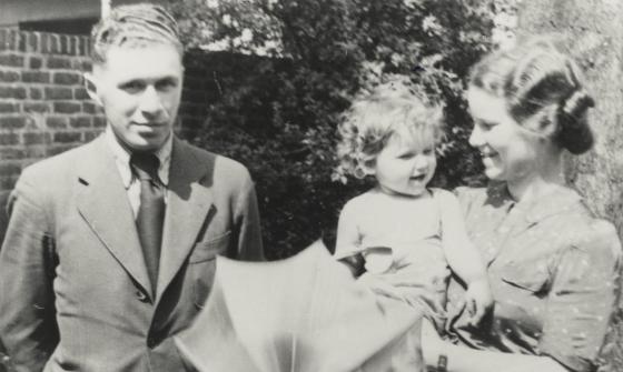 A black and white image of a man standing next to a woman holding a child and an umbrella.