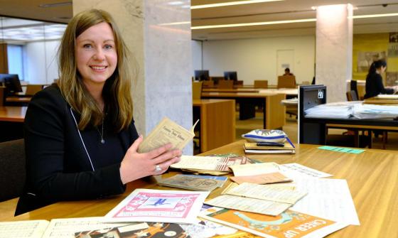 A person with long hair sitting at a table in a big room with lots of papers in front of them.