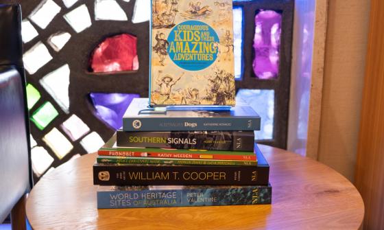 Stack of books on a round wooden table in front of colourful stained glass windows. The books are various sizes and colours