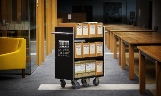 A National Library of Australia archival cart with labeled special collections boxes, placed in a reading room beside wooden tables and yellow seating.
