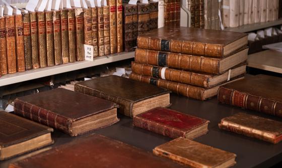 Shelf and table with dozens of old, leather-bound books, some with gold designs or writing on the spines