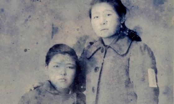 A black and white image of two women. One is seated and the other is standing with her arm around the seated woman.