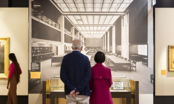 A tall man and shorter woman in the Treasures Gallery looking at a large black and white photo of the Library's main reading room taken in the 1960s. A woman in a pink top is looking at a framed artwork nearby.