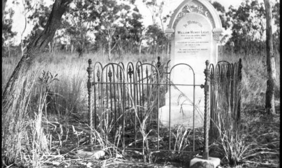 Black and white photograph of a gravestone surrounded by a wrought iron fence. The grave is situated in a grassy, rural setting with a few trees scattered around. The gravestone has an arched top and an inscription in memory of William Henry Light.
