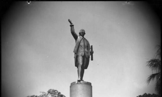 A black-and-white photograph of a statue of Captain Cook on a tall pedestal. The statue shows Cook standing with one arm raised and holding a rolled document in the other hand. Trees and part of a lamppost are visible in the foreground.