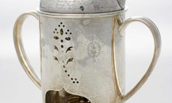 A silver polished kettle with ornate cutouts and inscription and printed crest. It has a tealight underneath.
