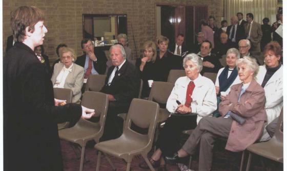A photo of a woman standing and speaking to a room of seated and standing people.