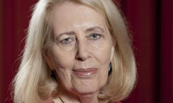 A colour photograph of Anne Summers. Anne has shoulder length blonde hair and wears a red outfit and a large round necklace, standing against a red curtain background.