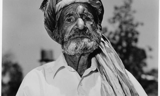 A black-and-white portrait of an older man with a weathered face and a beard, wearing a turban and a white shirt. He stands outdoors, with blurred trees in the background.