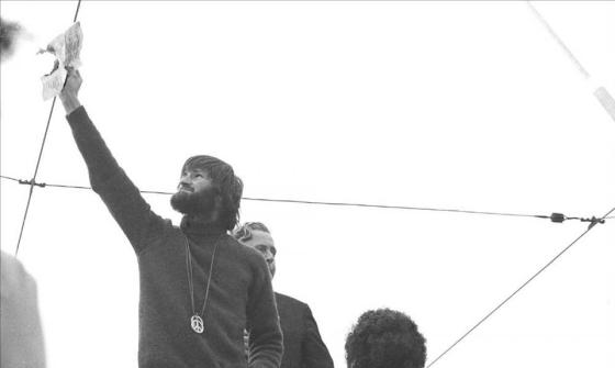 A black and white photo of a bearded man with collar length hair and a large metal peace medallion hanging down his front, holds a burning piece of paper up high, while several people watch and take photos.