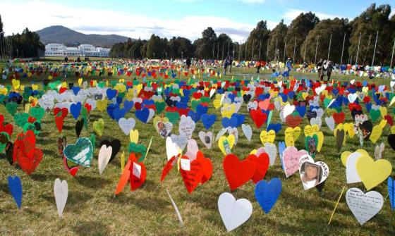 A large field filled with colorful heart-shaped signs, some with messages and images, displayed on the grass. Old Parliament House is visible in the background.