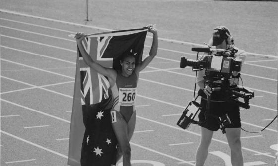Black and white photo of Cathy Freeman carrying Australian and Indigenous flags with a cameraman filming alongside her