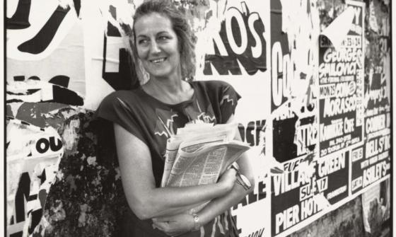 A black-and-white photograph of a woman standing against a wall covered in posters. She is smiling and holding a folded newspaper in her arms, casually dressed with loose hair. The posters in the background are partially torn and feature bold text and graphics.