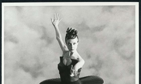 Ballerina Justine Summers strikes a pose for the camera. She is standing en point wearing a black tutu. She is standing infront of a textured curtain. She is staring directly at the camera.
