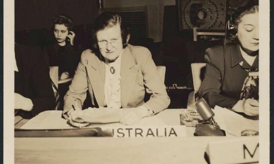 A black-and-white photograph of Jessie Street seated at a desk representing Australia at a United Nations conference. She is wearing a white shirt and suit jacket and is writing, with other women visible beside her.