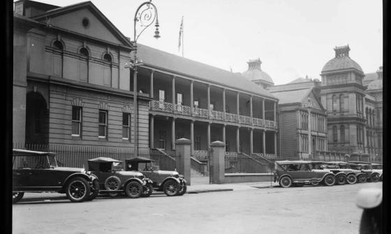 black and white photo of old buildings with old cars in the street out the front