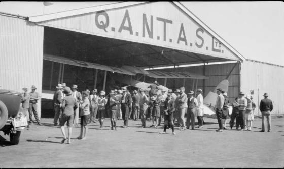 Black and white photo of a crowd of people examining a biplane in a QANTAS hanger