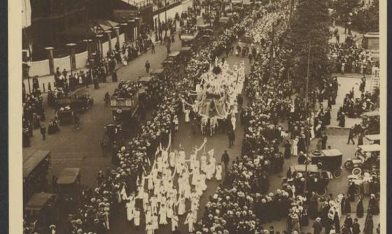 An aerial view of a large street procession with participants dressed in white, marching in rows and carrying banners. The crowded scene is framed by towering buildings on one side and tree-lined streets on the other. Spectators line the sidewalks, watching the organised march as cars and horse-drawn carriages are scattered among the crowd. 