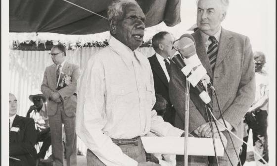 Vincent Lingiari addresses a crowd at a microphone while Gough Whitlam stands beside him. Several people are visible in the background, observing.