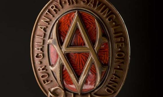 An oval shaped bronze medal. The oval is made of a belt buckled at the bottom. At the top is a stylised royal crown. In the centre of the oval is a monogram made up of an intertwined "V.A." Behind the monogram is pattered red glass. Around the oval in raised letters are the words 'For gallantry in saving life on land'. The medal is held by a ribbon of red and white vertical stripes